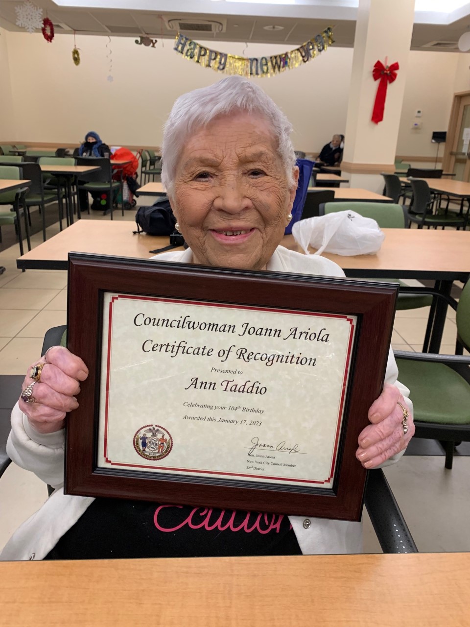 Woman is seated at a table holding a framed Certificate of Recognition.